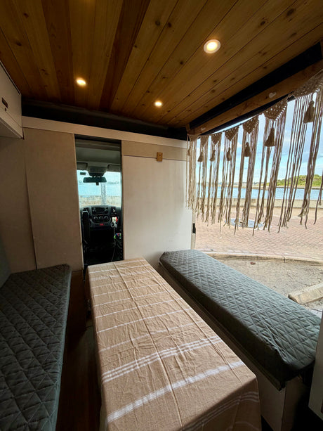 Cozy RV rental interior with wooden ceiling, quilted beds/benches, macrame curtain, and a scenic waterfront view outside.