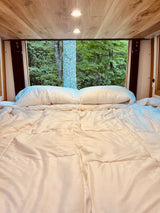Enjoy a serene forest view from the cozy bed of this RV rental, featuring white bedding and a warm wooden ceiling with lights.
