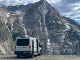 Adventure awaits! This white RV rental is parked among stunning, snow-capped mountains under a cloudy sky.