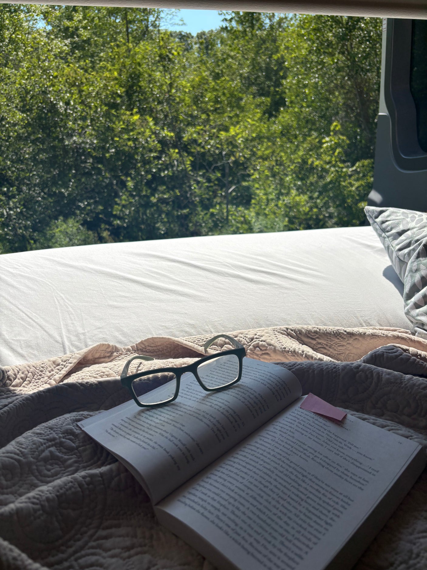 Cozy reading in an RV rental! A book and glasses on a comfy bed, with a serene view of green trees outside.