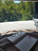 Cozy reading in an RV rental! A book and glasses on a comfy bed, with a serene view of green trees outside.