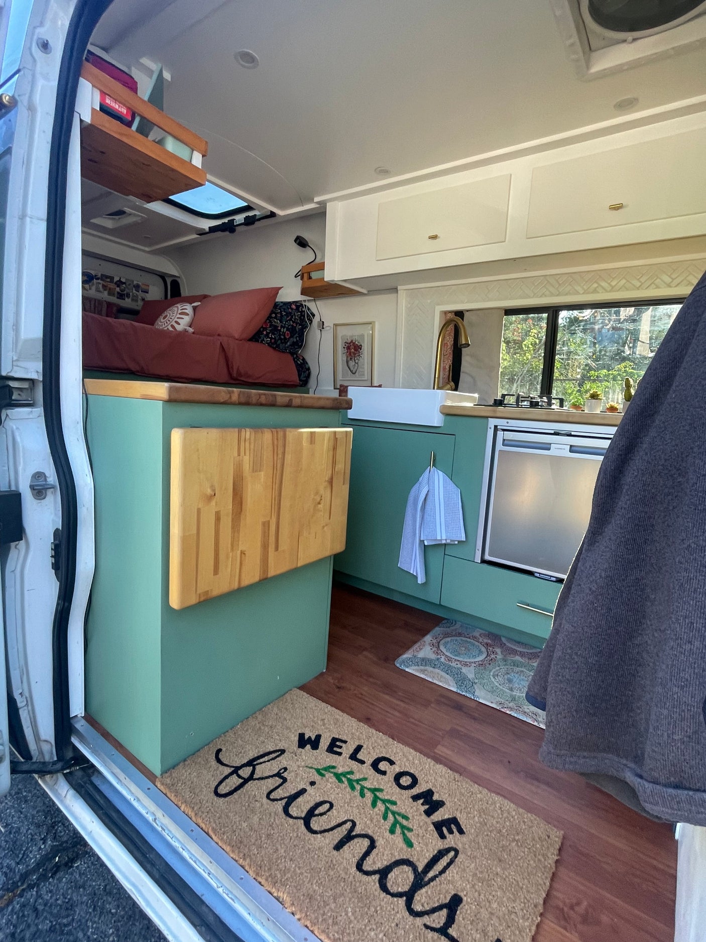 Cozy rv rental interior: green kitchen, comfy bed, and 'WELCOME friends' mat. Perfect compact living space.