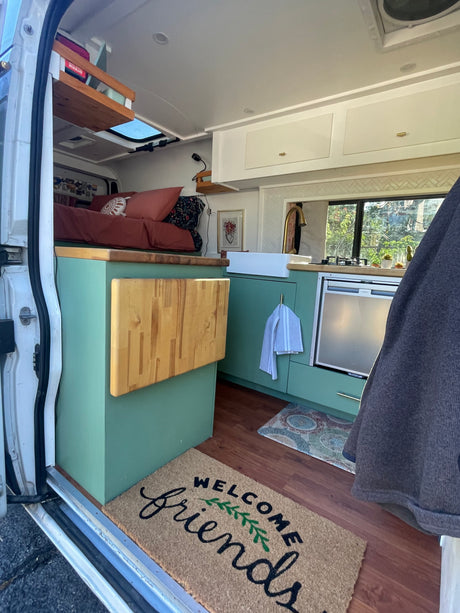 Cozy rv rental interior: green kitchen, comfy bed, and 'WELCOME friends' mat. Perfect compact living space.