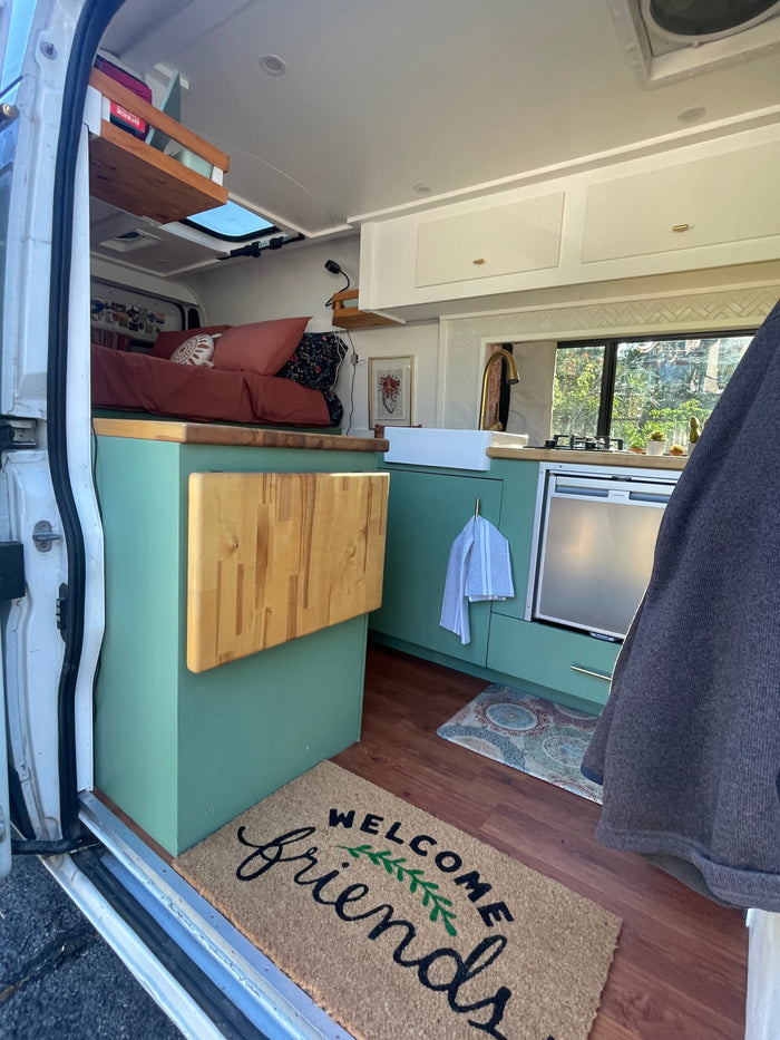 Cozy rv rental interior: green kitchen, comfy bed, and 'WELCOME friends' mat. Perfect compact living space.