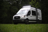 White Ford Transit camper van with bright roof lights & off-road tires, parked on grass. Ideal for your next rv rental adventure.