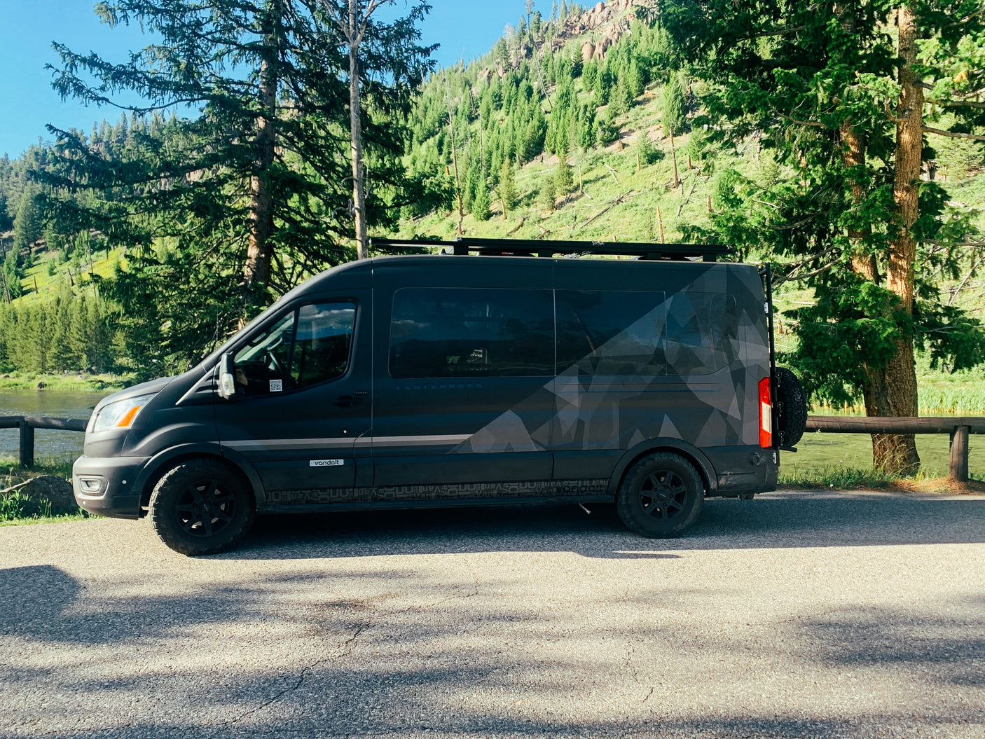 Discover nature with this dark gray adventure van, an ideal RV rental, by a river and green mountains.