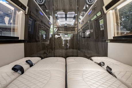 Luxury RV rental: plush quilted beds with seatbelts, glossy wood walls, windows, and central TV offer travel comfort.
