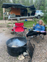 Enjoying a cozy RV rental campsite. Woman, dog, teardrop camper, & SUV relax by the fire.