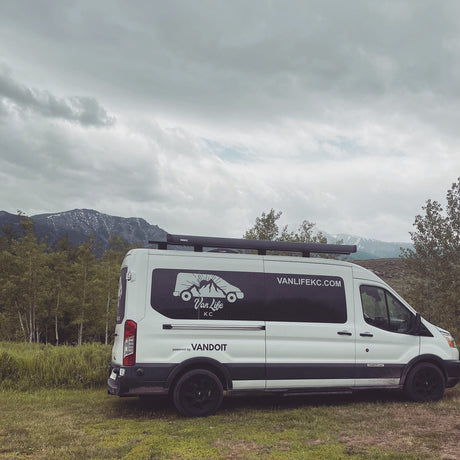 A white RV rental van with black graphics is parked on green grass, backed by trees, snow-capped mountains, and a cloudy sky.