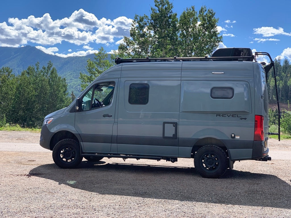 Gray Winnebago Revel 4x4 rv rental parked on gravel with mountains and trees under a blue sky.