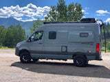 Gray Winnebago Revel 4x4 rv rental parked on gravel with mountains and trees under a blue sky.