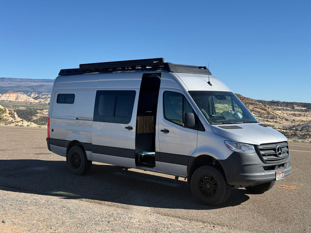 Silver Mercedes Sprinter camper van, an RV rental, parked with its door open revealing the interior, against a scenic desert landscape.