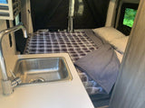 Compact RV rental interior features a cozy bed with plaid bedding and a small kitchen sink, ideal for travel.