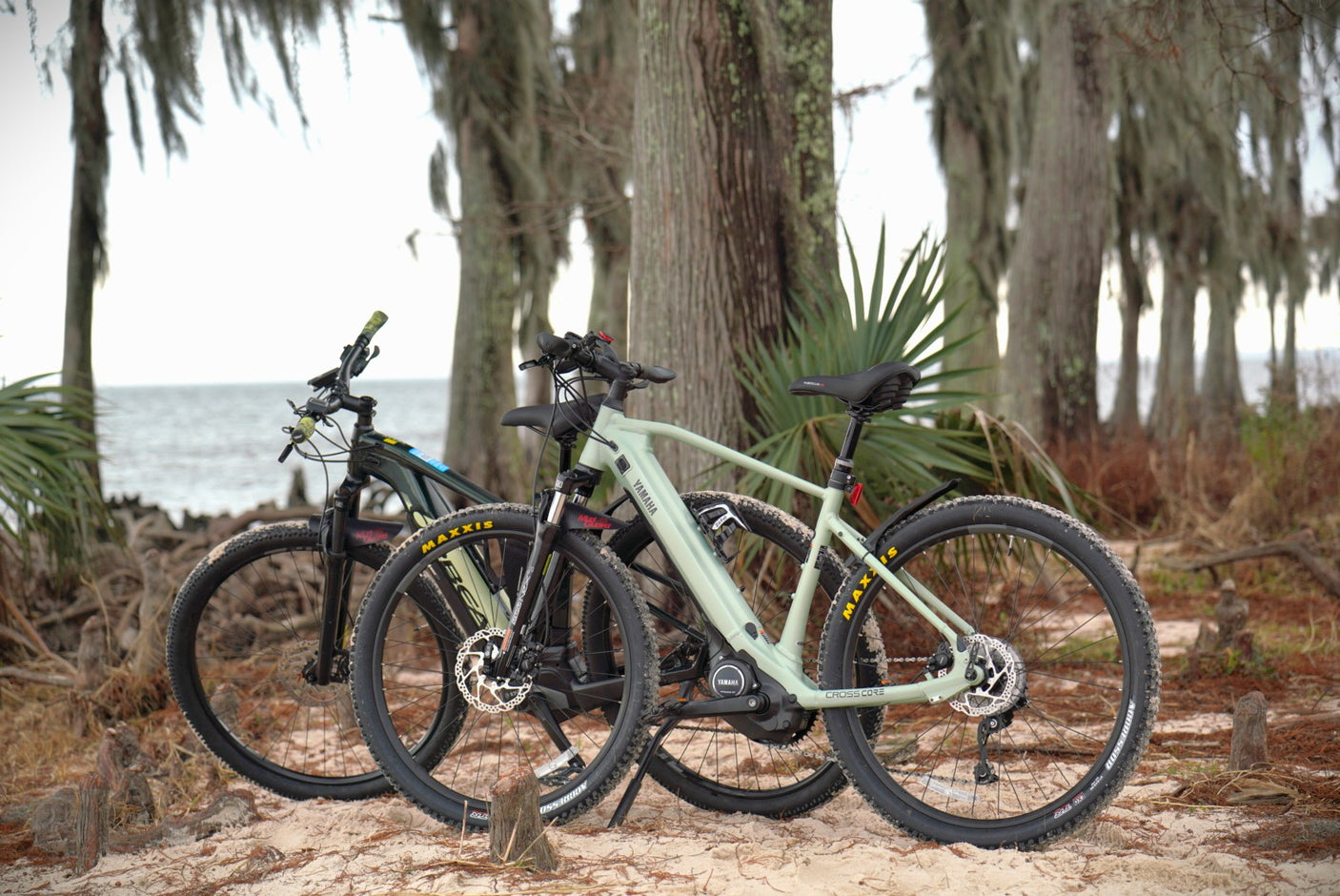 Two Yamaha e-bikes on a sandy path near water and trees. Perfect for exploring on your next RV rental adventure!