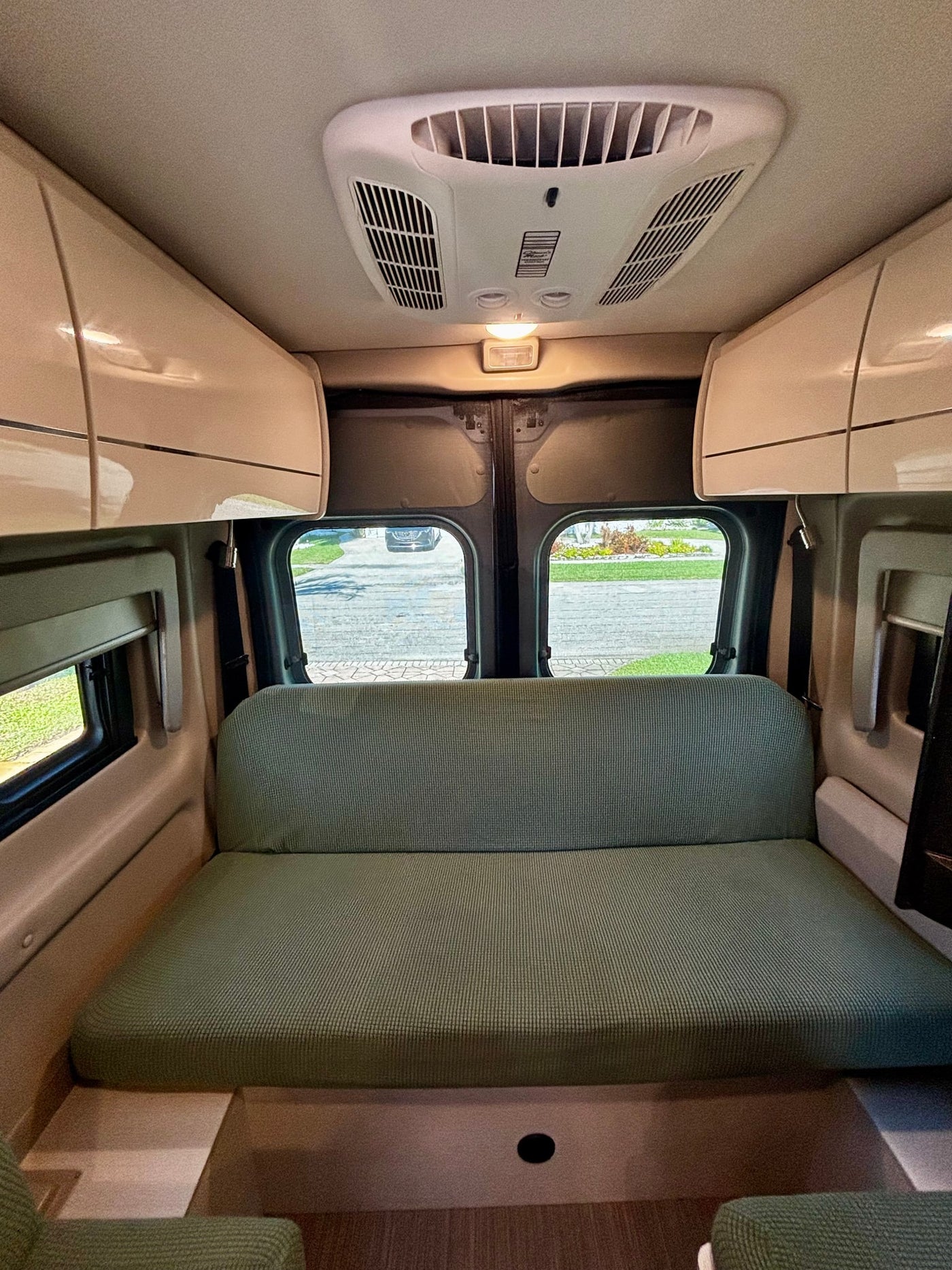 This RV rental interior features a green sofa/bed, overhead storage, an AC unit, and rear windows showing an outside view. — photo 17