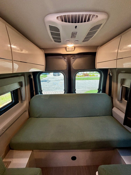 This RV rental interior features a green sofa/bed, overhead storage, an AC unit, and rear windows showing an outside view.