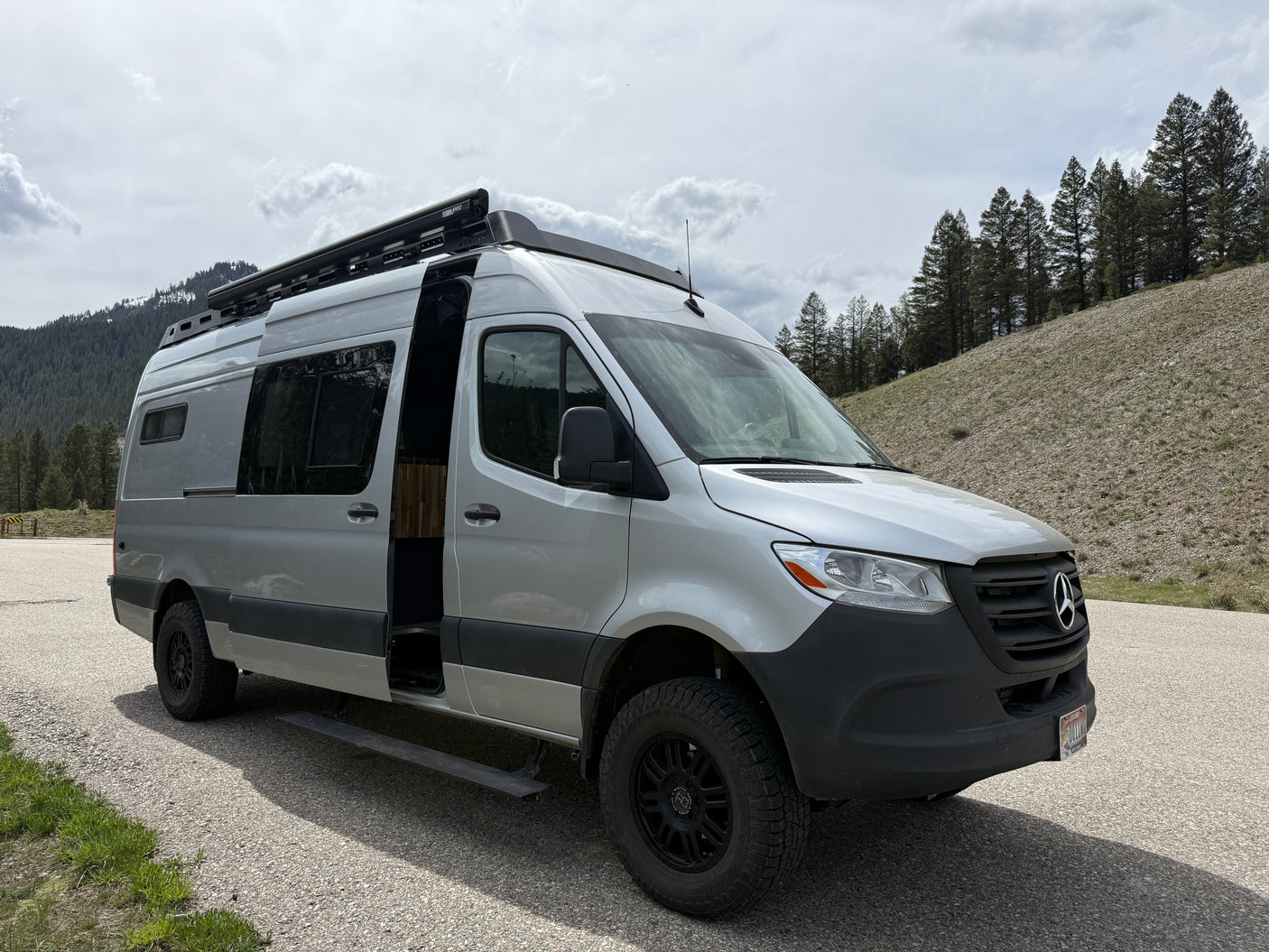 Silver Mercedes Sprinter camper van RV rental with an open door, parked outdoors near mountains and trees.