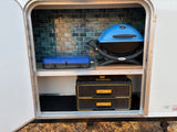 RV rental adventure: Compact outdoor kitchen with blue portable grill, camping stove, and DeWalt storage in a tiled bay.