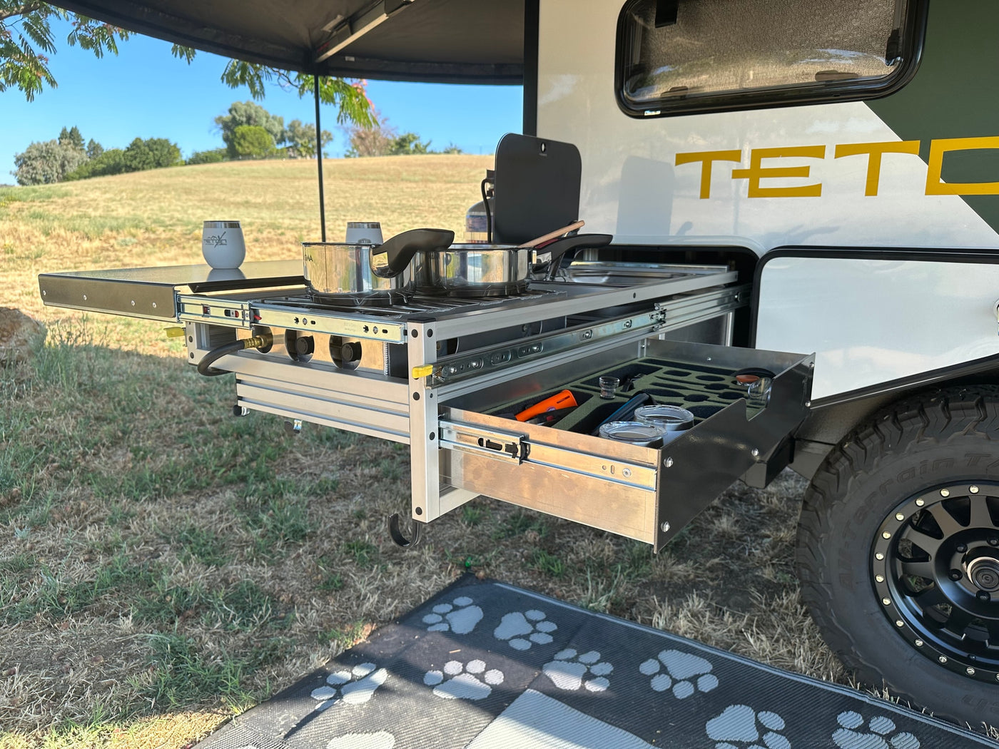 Rv rental outdoor kitchen: a pull-out stove with pots, and a drawer of utensils, set up for cooking amidst grassy hills. — photo 7