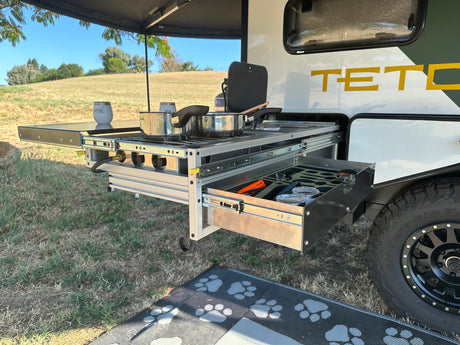 Rv rental outdoor kitchen: a pull-out stove with pots, and a drawer of utensils, set up for cooking amidst grassy hills.