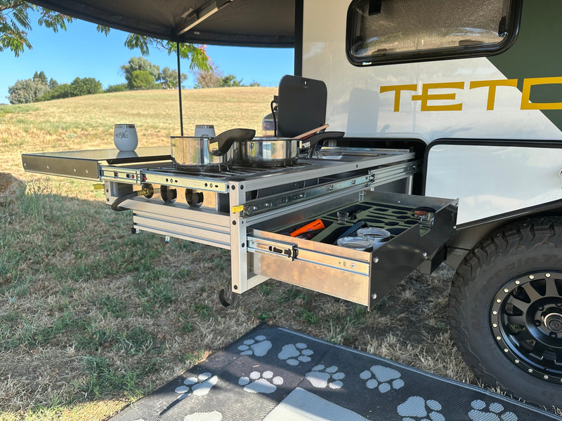 Rv rental outdoor kitchen: a pull-out stove with pots, and a drawer of utensils, set up for cooking amidst grassy hills. — photo 7