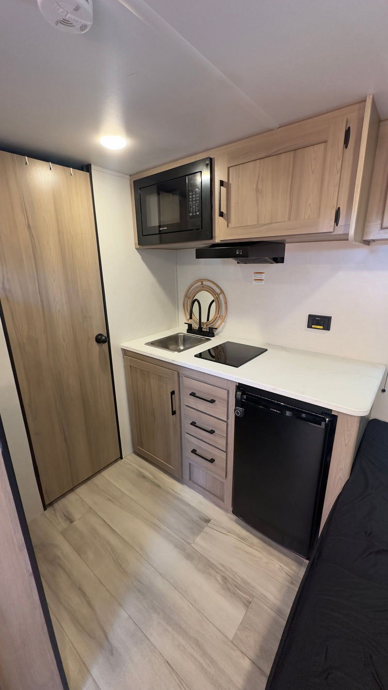 Modern RV rental kitchenette: light wood cabinets, black microwave, sink, cooktop, and compact fridge are visible. — photo 6