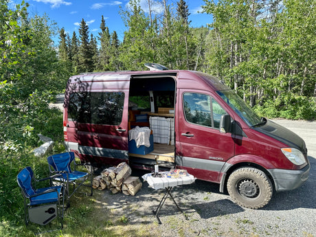 Enjoying the outdoors with a maroon RV rental. Campervan parked with open door, chairs, firewood, and a table ready for a meal.