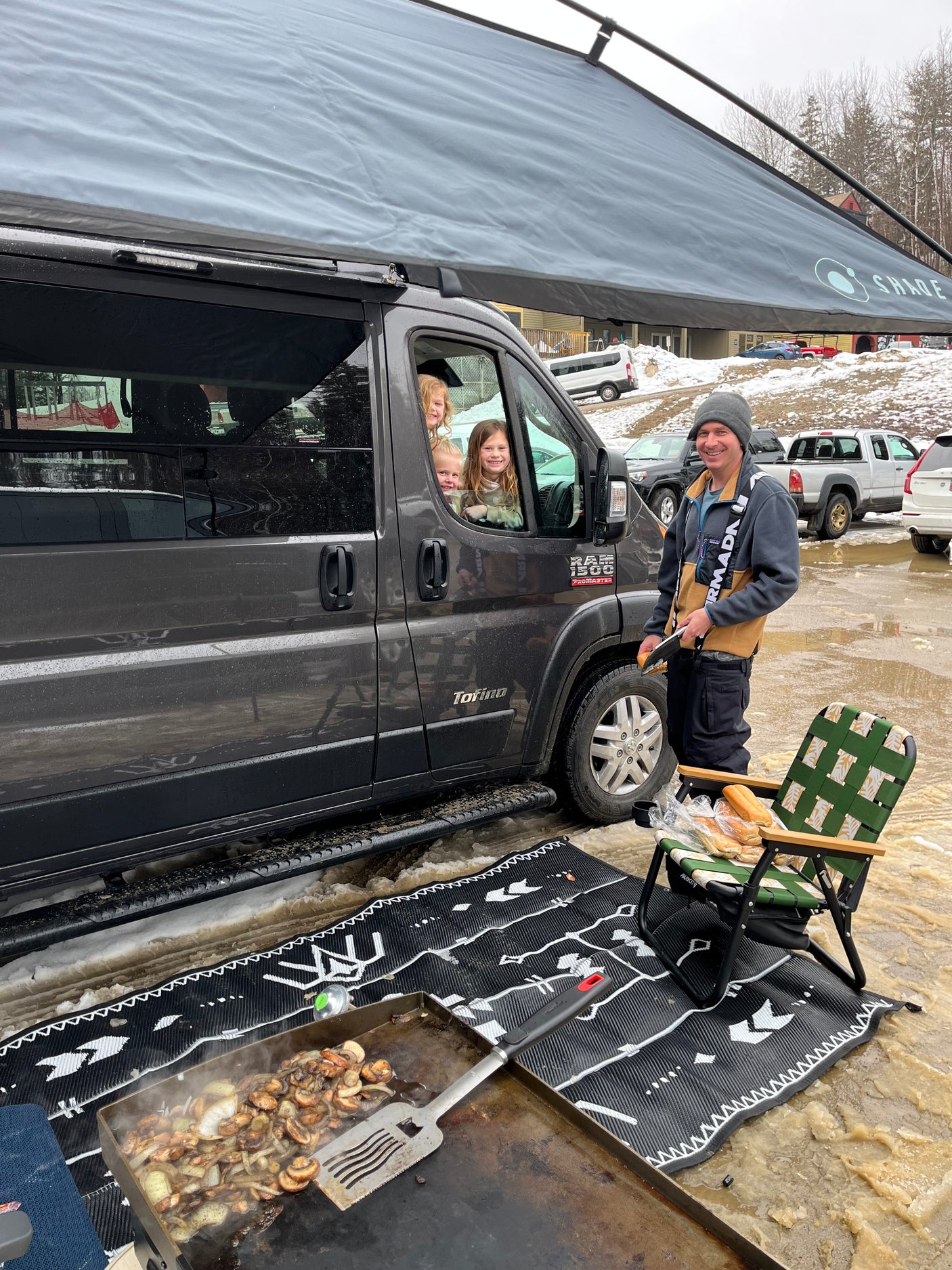 RV rental life: Kids in the van, man cooking on a griddle outside under an awning on a muddy/snowy day.