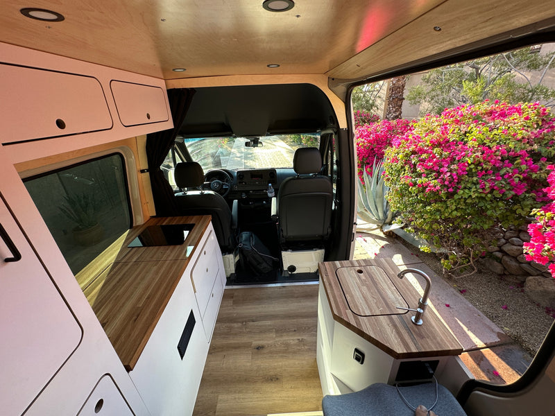 Spacious RV rental interior featuring a modern kitchen, wooden accents, and an open door view of lush plants outside. Perfect for van life! — photo 9