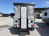 A gray Clipper pop-up RV rental, seen from the rear with its door open, parked in a sunny lot surrounded by other RVs.