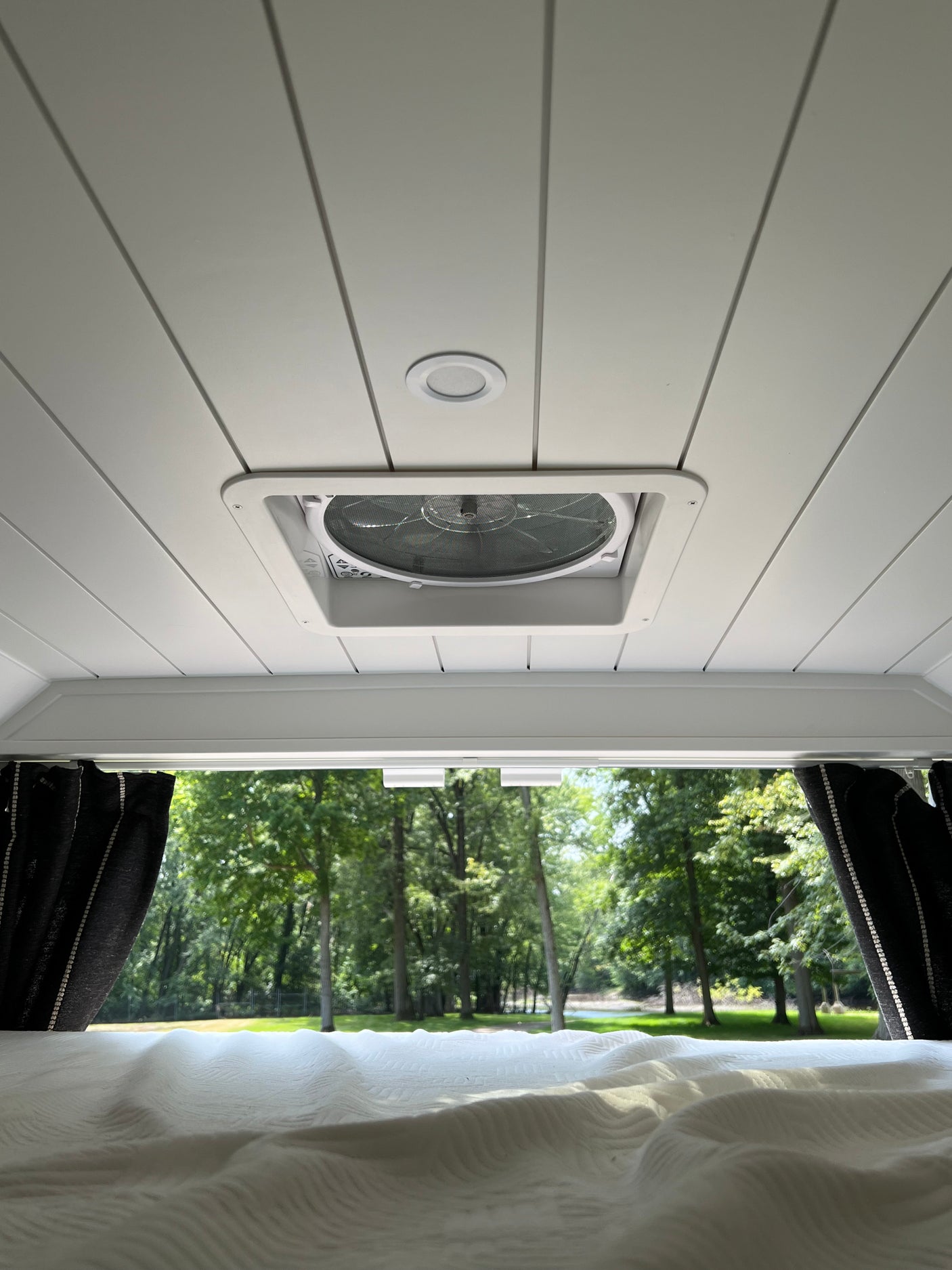 Serene RV rental interior: white ceiling with vent fan and light, a comfy bed, and a scenic view of lush trees outside.