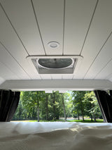 Serene RV rental interior: white ceiling with vent fan and light, a comfy bed, and a scenic view of lush trees outside.