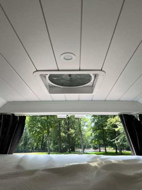 Serene RV rental interior: white ceiling with vent fan and light, a comfy bed, and a scenic view of lush trees outside.