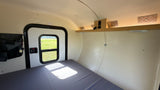 Compact rv rental interior with a bed, exit door, and overhead shelf with controls. Ready for your adventure.