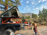 Truck with rooftop tent for camping. Cooking outdoors, an alternative to RV rental, amidst hills and pines.