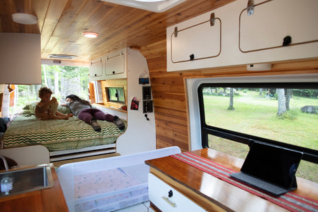 Cozy RV rental interior: a woman & child relax on a bed amidst wooden walls, a kitchen, and a laptop with outdoor views.