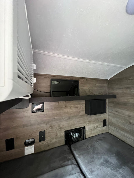 View of an RV rental interior with wood-grain walls, showcasing an overhead AC unit, wall-mounted TV, control panel, and beds.