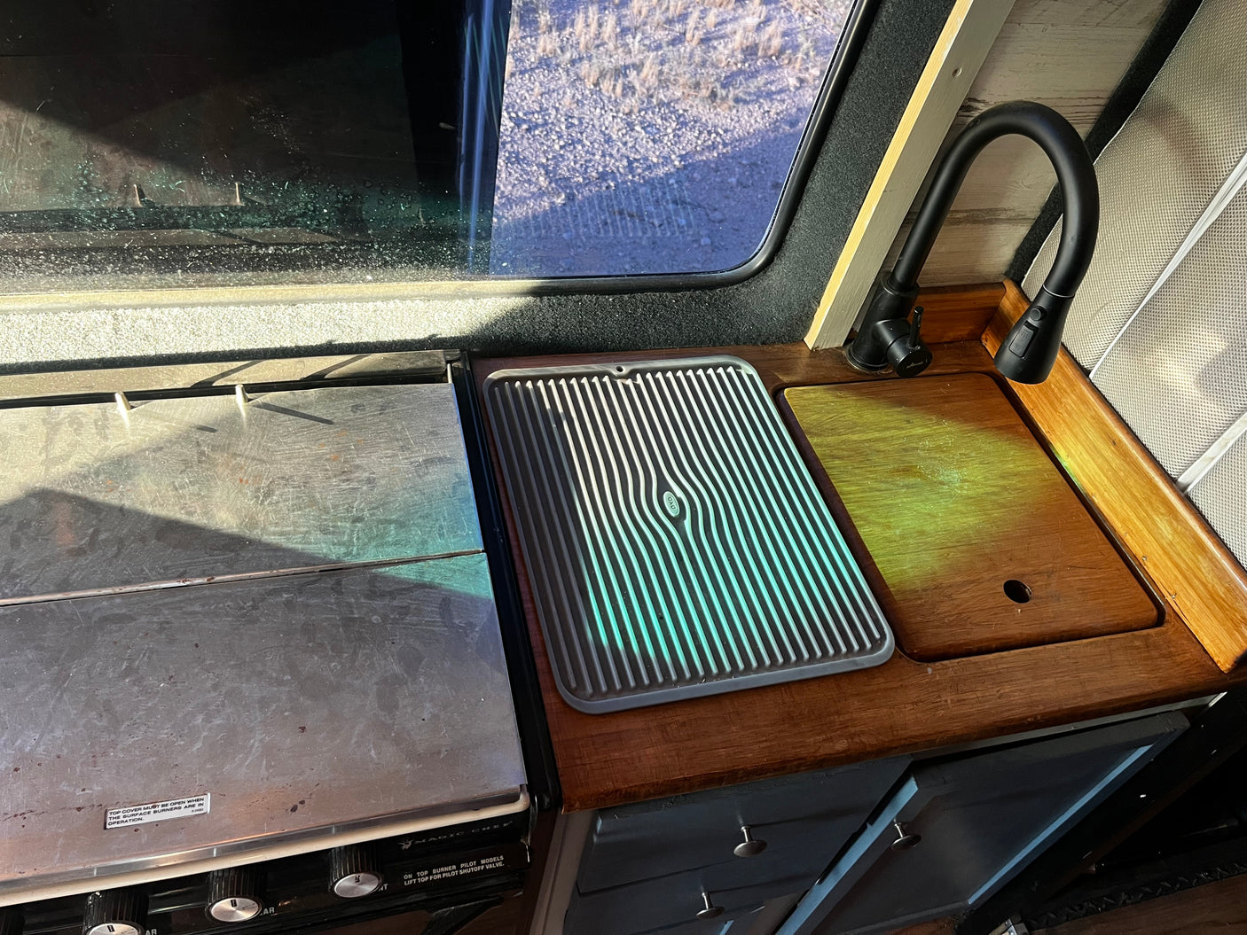 RV rental galley showing stove, sink with wooden cover, flexible faucet, OXO drainer, and outdoor window view.
