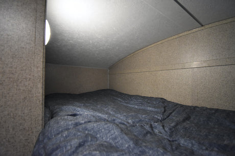Cozy RV rental bunk with a blue comforter, lit by a side lamp under a curved ceiling.