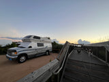 Silver RV rental camper van with sunshades parked on a dirt lot. Two dogs are on a wooden boardwalk under a sunset sky.