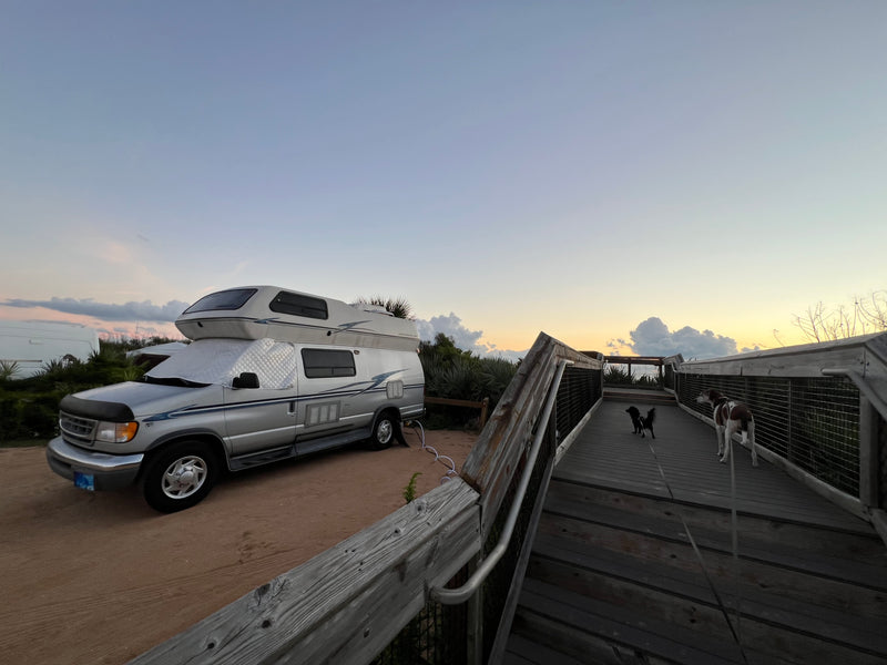 Silver RV rental camper van with sunshades parked on a dirt lot. Two dogs are on a wooden boardwalk under a sunset sky. — photo 12