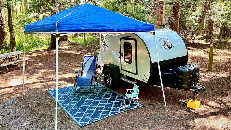 Cozy RV rental setup: a white teardrop camper, blue canopy, chairs, and rug at a peaceful wooded campsite. — photo 16