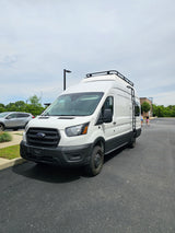 This white Ford Transit camper van, likely an RV rental, features a roof rack & side ladder.