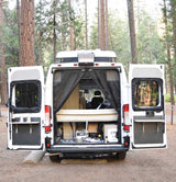 Rear view of a white RV rental camper van with open doors, revealing its interior bed, storage, and person inside. Great for camping.