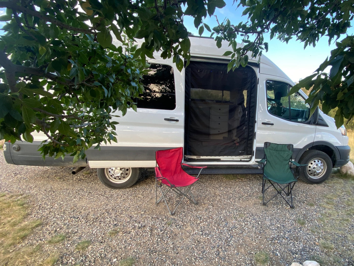  set against a stunning gradient sunset sky.|RV rental: white camper van with open side door (screen) and two folding chairs on gravel under trees.|Cozy bed inside an RV rental with a stunning view of a golden field and distant hills at sunrise.|RV rental kitchen: Cooking pot with spoon — photo 2