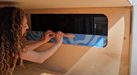 Customizing an RV rental interior: A person attaches a decorative panel below a window in a wooden camper.