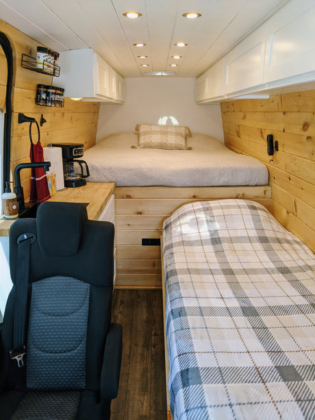 Cozy RV rental interior with elevated main bed, side bunk, compact kitchen, and warm wood finishes.