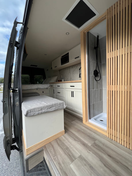 Modern RV rental interior featuring a white kitchen, a comfortable bed, and a sleek, tiled shower with wood accents.