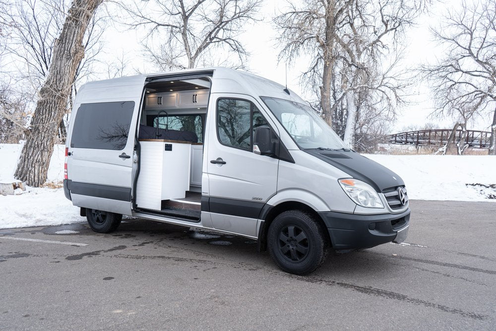RV rental: Silver Mercedes Sprinter camper van with open side door showing interior, parked in winter snow.