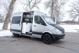 RV rental: Silver Mercedes Sprinter camper van with open side door showing interior, parked in winter snow.